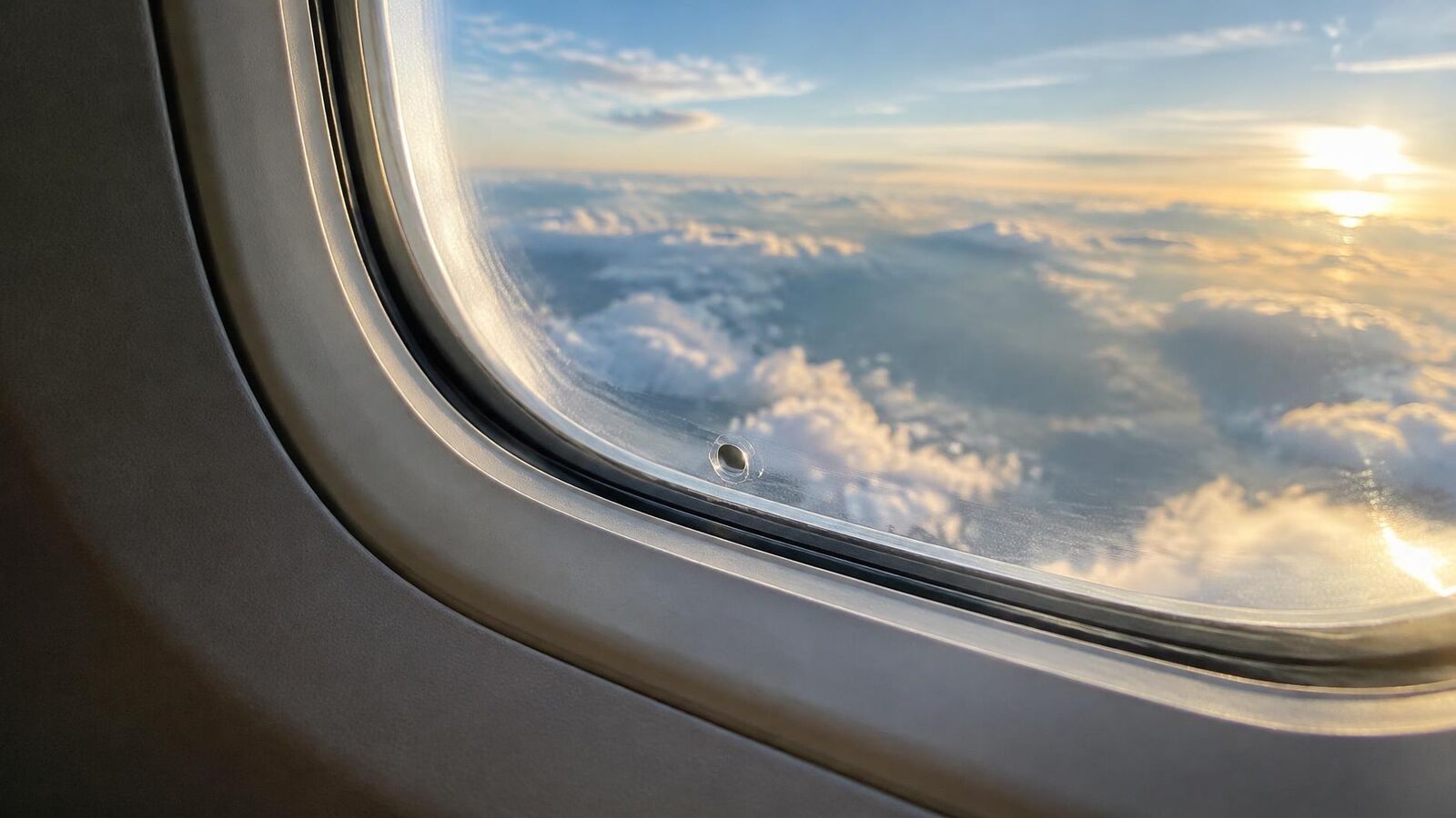 Janela de avião com o pequeno furo no painel interno e nuvens ao fundo